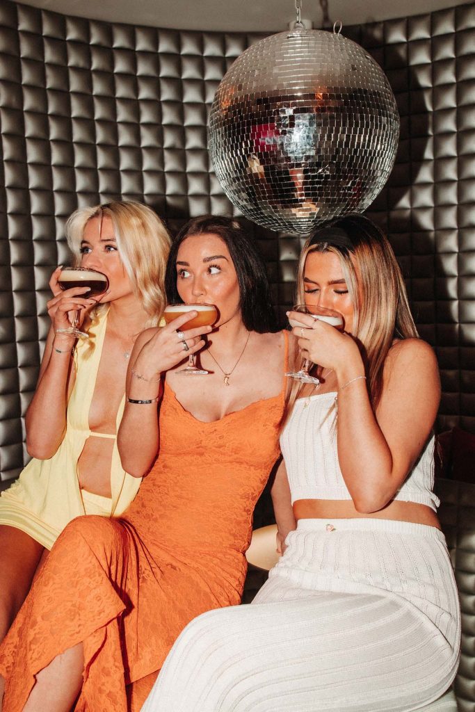 three girls drinking cocktails under a disco ball